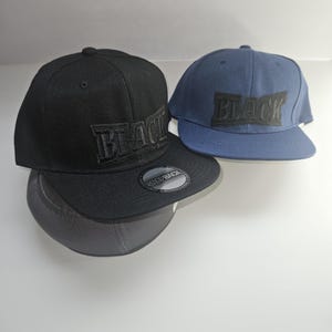 May include: A black snapback cap and a blue snapback cap, both featuring the word BLACK embroidered in a bold, block font on the front panels. The black cap includes a circular sticker on the flat brim that reads SNAPBACK in white text.