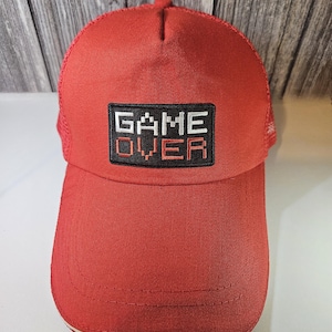 May include: Red and black trucker hat with a black patch that says "GAME OVER" in white and red pixelated letters.