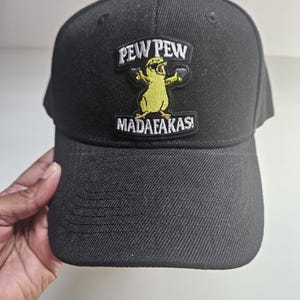 May include: A black baseball cap featuring an embroidered patch on the front. The patch depicts a yellow cartoon chick wearing sunglasses and holding two small black guns, with the text PEW PEW MADAFAKAS! embroidered in white and black.