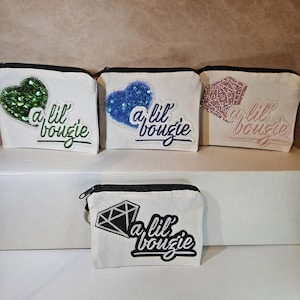 Puede incluir: Monederos de lona blanca con cremalleras negras. Cada monedero presenta un diseño de lentejuelas: un corazón verde, un corazón azul, un diamante rosa y un diamante negro. La frase "a lil bougie" está bordada en cada monedero.
