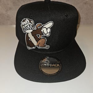 Könnte beinhalten: Schwarze Snapback-Kappe mit einem braunen und weißen Cartoon-Affen-Patch. Der Affe zeigt mit einem wütenden Gesichtsausdruck. Die Kappe hat einen flachen Schirm und einen Schnappverschluss. Aufkleber "Snapback".