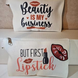May include: Two white canvas cosmetic bags. One bag has the text "Beauty is my business" with a lip graphic and mascara brush. The other bag has the text "But first Lipstick" with a lipstick and lip graphic.