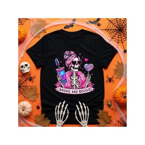 May include: A black t-shirt featuring a graphic of a skeleton with pink hair, heart-shaped sunglasses, and a pink fur coat. The skeleton holds a galaxy-patterned tumbler and a heart-shaped lollipop. Below the graphic is the text BROKE AND BOUGIE.