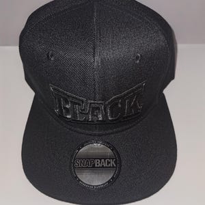 May include: A black snapback hat with the word "BLACK" embroidered on the front. The hat has a flat brim and a structured crown. The hat also has a "SNAPBACK" sticker on the brim.