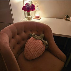 Puede incluir: Un cojín de fresa rosa tejido a ganchillo, con hojas verdes, descansa sobre un sillón de terciopelo rosa. El cojín es el punto focal, con una apariencia texturizada. El fondo incluye un escritorio, un jarrón de rosas rojas y una pequeña lámpara.
