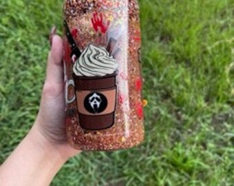 May include: A clear, glitter-filled tumbler with a brown coffee cup graphic and a white whipped cream design. The tumbler also features a red handprint and a ghost face graphic. The tumbler is held by a person.