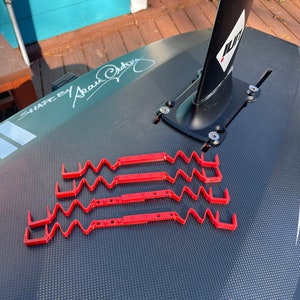 May include: Four red plastic straps with a wavy design are attached to a black carbon fiber surfboard. The straps are used to secure a surfboard fin to the board. The text "SHAPED BY Sean Ordonez" is visible on the board.