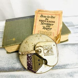 May include: A vintage circular metal Kriss Kross razor stropper with a manual crank handle, featuring a worn metallic finish and embossed branding. Beside it lies a small, aged paper instruction booklet titled How to Use the Kriss Kross Stropper and Razor.