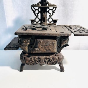 May include: Antique cast iron miniature stove with ornate detailing. Features a small chimney, side panels, and the word "CRESCENT" embossed on the front. The stove has a rustic, aged appearance, suggesting it is a vintage item.