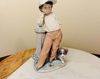 May include: A porcelain figurine of a boy wearing a pink and white baseball cap, leaning against a stone pillar. He is wearing a white shirt, brown pants, and brown shoes. A small brown and white dog is standing next to him. The figurine is on a white marble surface.