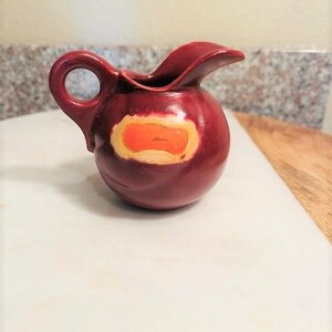 May include: A small, red ceramic creamer with a yellow and orange design. The creamer has a rounded body and a small handle.