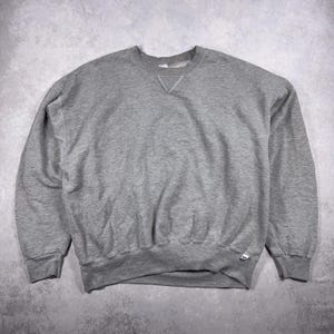 May include: A heather gray crew neck sweatshirt. The sweatshirt has a classic design with a ribbed collar, cuffs, and hem. The garment is made of a soft, comfortable material, perfect for casual wear. The Russell Athletic logo is visible on the bottom hem.