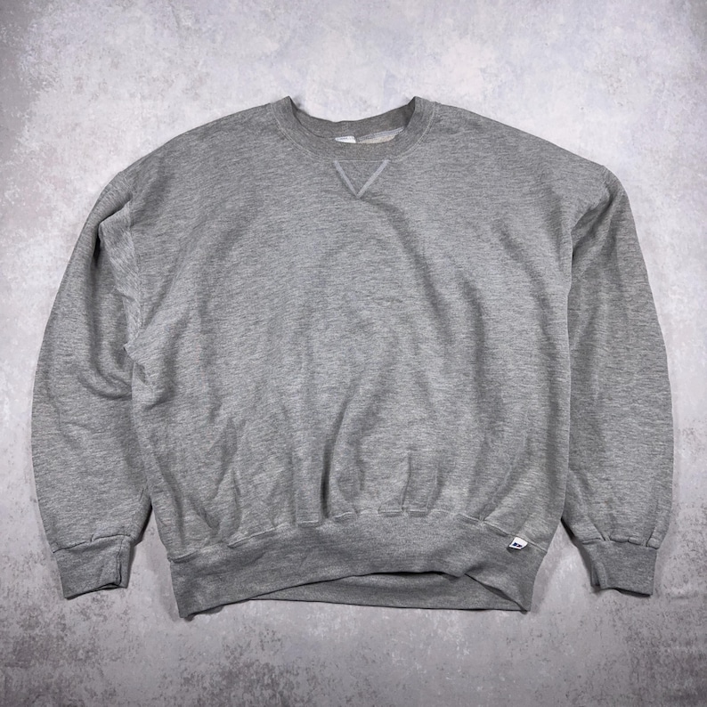 May include: A heather gray crew neck sweatshirt. The sweatshirt has a classic design with a ribbed collar, cuffs, and hem. The garment is made of a soft, comfortable material, perfect for casual wear. The Russell Athletic logo is visible on the bottom hem.
