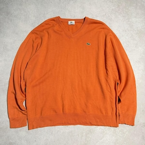 May include: Orange V-neck sweater with long sleeves. The sweater has a small green crocodile logo on the chest and a small white tag near the neckline. The sweater is made of a soft material.