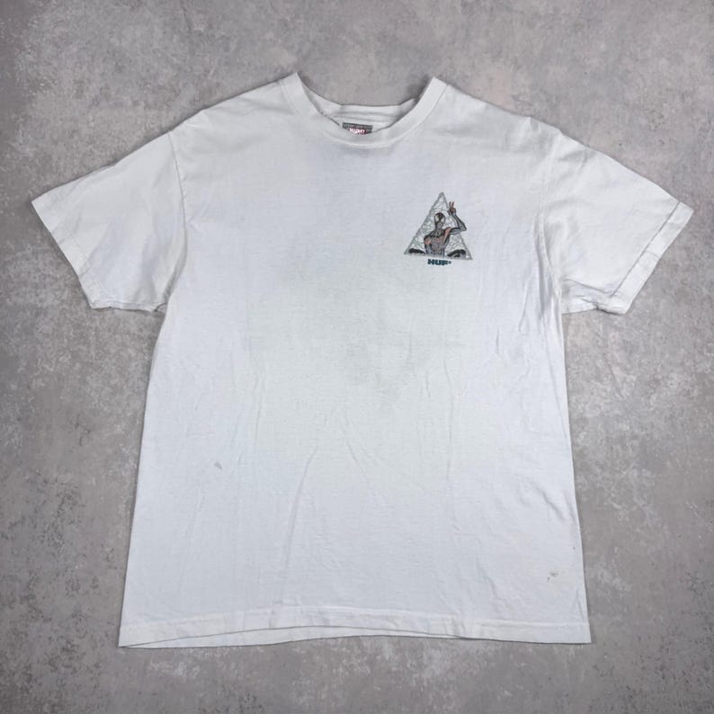 May include: White t-shirt with a small graphic on the front. The graphic is a triangle with a figure inside, making a peace sign. The word "HUF" is below the graphic. The shirt is made of a soft material.