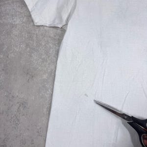 May include: A white t-shirt with a slightly wrinkled texture. A pair of black scissors with silver blades is in the lower right corner. The shirt is on a gray surface.