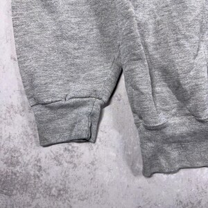 May include: Close-up of a gray sweatshirt sleeve and cuff. The fabric has a textured appearance. The cuff is ribbed and the sleeve is a solid color. The background is a mottled gray.
