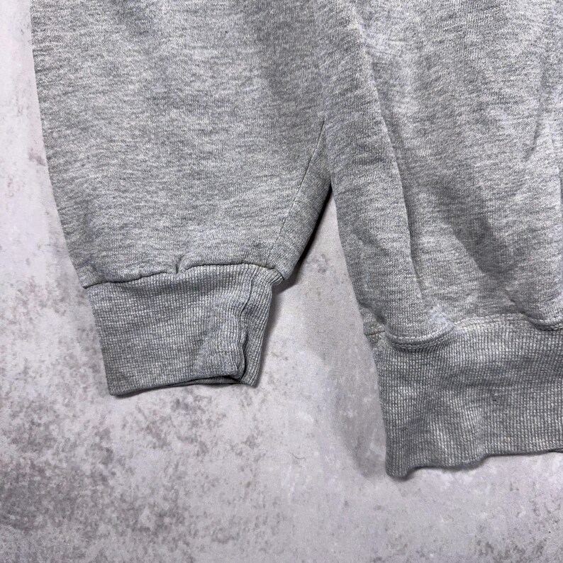 May include: Close-up of a gray sweatshirt sleeve and cuff. The fabric has a textured appearance. The cuff is ribbed and the sleeve is a solid color. The background is a mottled gray.