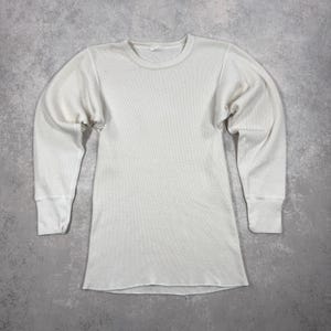 May include: Off-white thermal long-sleeved shirt with a crew neck and ribbed cuffs. The shirt has a waffle-knit texture and a relaxed fit. The garment is laid flat on a grey surface.