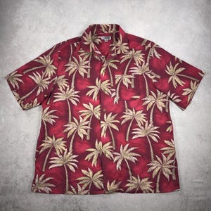 Puede incluir: Camisa de manga corta con fondo rojo y un estampado repetido de palmeras beige. La camisa tiene cuello y un bolsillo en el pecho izquierdo. La tela parece ser un material ligero.