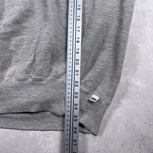 May include: A gray sweatshirt with a ribbed hem and cuffs. A ruler is placed next to the garment, showing measurements in inches. A small white tag with a blue and red logo is visible on the cuff.