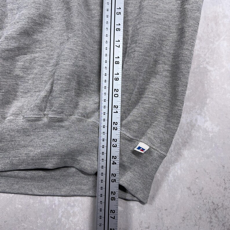 May include: A gray sweatshirt with a ribbed hem and cuffs. A ruler is placed next to the garment, showing measurements in inches. A small white tag with a blue and red logo is visible on the cuff.