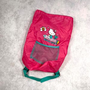 Puede incluir: Una mochila rosa brillante con cordón, con un diseño de Hello Kitty, una abeja y una rana. La mochila tiene un bolsillo de malla turquesa y correas turquesas. Las palabras "HELLO KITTY" están impresas en el lápiz.