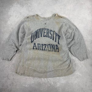 May include: A vintage gray sweatshirt with the words "UNIVERSITY ARIZONA" printed in navy blue. The sweatshirt shows signs of wear, including holes and discoloration, suggesting it is a well-worn item. The sleeves are three-quarter length.