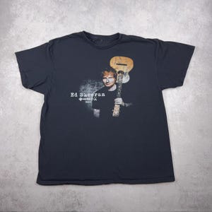Peut inclure: T-shirt noir avec un graphisme d'Ed Sheeran tenant une guitare. Le texte "Ed Sheeran" et "2008" sont imprimés en blanc. Le t-shirt est de style col rond.