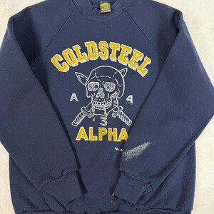 May include: Navy blue sweatshirt with the text "COLDSTEEL" in yellow block letters above a skull graphic with crossed knives. The graphic also includes the word "ALPHA" and the numbers "A 4 3 4".