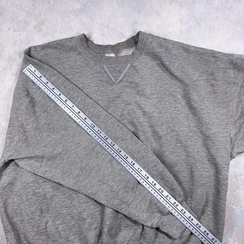 May include: A heather gray sweatshirt with a crew neck and a V-shaped detail at the neckline. A ruler is placed across the sleeve, measuring approximately 27 inches. The sweatshirt is made of a soft, comfortable material.