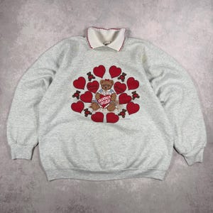 May include: A light gray sweatshirt with a white collar and red trim. The front features a design of red hearts and teddy bears surrounding a teddy bear holding a heart that says "Grandma's Lovelies".