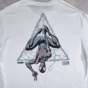 May include: White t-shirt featuring a graphic of a superhero in a black and gray suit hanging upside down within a gray web design. The shirt has the "HUF" and "MARVEL" logos.