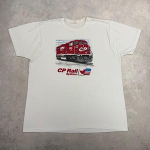 以下が含まれることがあります： 赤いCP Rail Systemの電車のグラフィックが特徴の白いTシャツ。グラフィックには、CP Railのロゴ、カナダのメープルリーフ、アメリカの国旗が含まれています。シャツはクルーネックで半袖です。