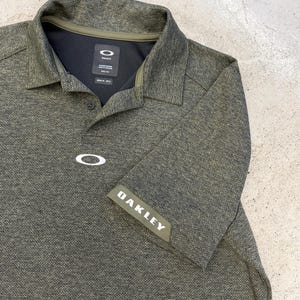 May include: Olive green polo shirt with the Oakley logo on the chest and sleeve. The shirt has a textured pattern, a black collar, and a button-down placket. The Oakley logo is white.