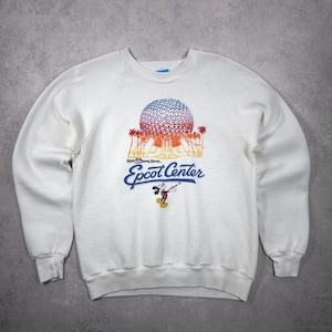 Puede incluir: Sudadera blanca de cuello redondo con un gráfico colorido de la bola del Centro Epcot, palmeras y Mickey Mouse. El texto "Epcot Center" está impreso en azul. La sudadera tiene mangas largas y puños acanalados.