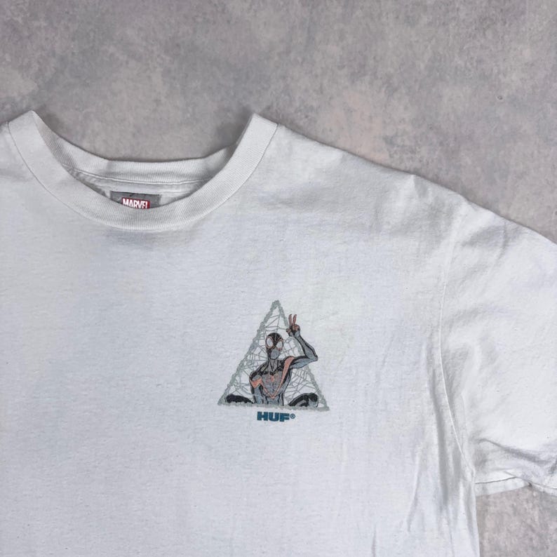 May include: White t-shirt featuring a Marvel logo and a graphic of Spider-Man inside a triangle, with the brand name "HUF" below. The Spider-Man graphic is in shades of gray, red, and black.