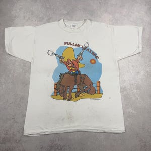 Pode incluir: Camiseta branca com um desenho de um cowboy de desenho animado montando um cavalo selvagem. O cowboy está segurando duas armas e a camisa tem o texto "Pullin' Leather". O design inclui um chapéu amarelo, céu azul e um cavalo marrom.