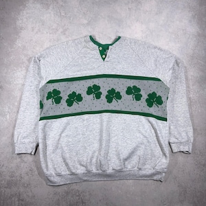 May include: A gray sweatshirt with a green and gray shamrock design across the chest. The sweatshirt has a green collar and three buttons. The long-sleeved top is made of a soft material.