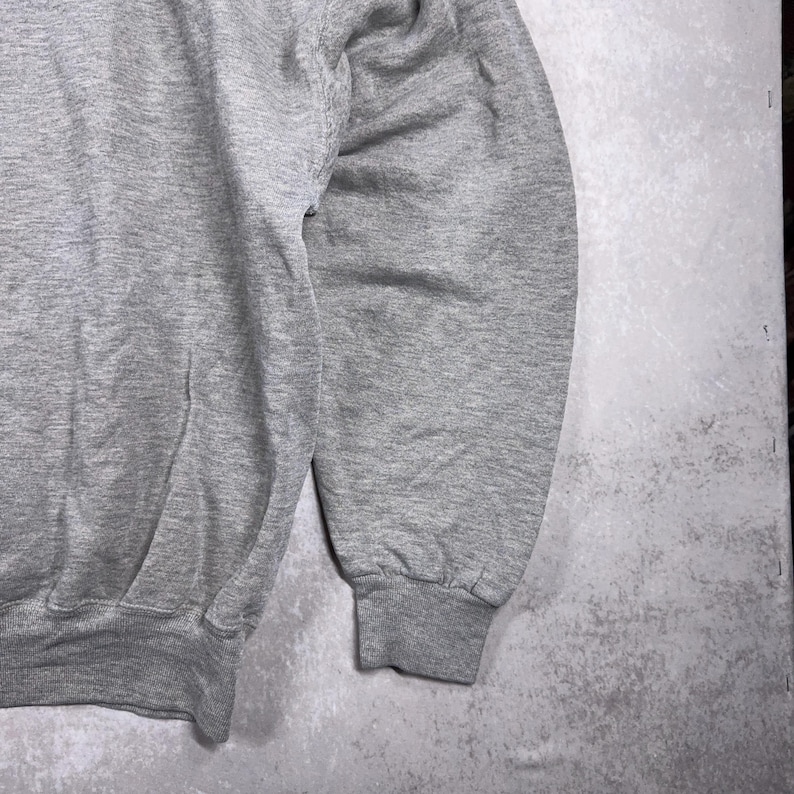 May include: Close-up of a heather gray sweatshirt. The image shows the sleeve and part of the body of the garment. The cuff is ribbed, and the fabric appears to be a soft, comfortable material. The background is a mottled gray.