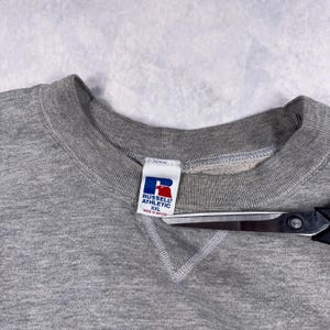 May include: A heather gray Russell Athletic sweatshirt with a crew neck. The tag features the brand logo in blue and red, with the text "RUSSELL ATHLETIC" and "XXL Made in Mexico". A pair of scissors is partially visible.