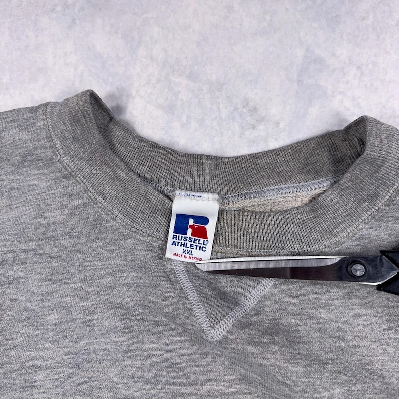 May include: A heather gray Russell Athletic sweatshirt with a crew neck. The tag features the brand logo in blue and red, with the text "RUSSELL ATHLETIC" and "XXL Made in Mexico". A pair of scissors is partially visible.