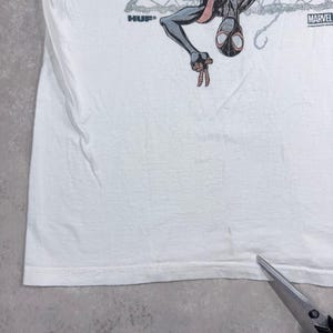 May include: White t-shirt featuring a graphic of Spider-Man in black, red, and gray. The shirt has the "HUF" logo on the left and the "MARVEL" logo on the right. The t-shirt is made of a soft, comfortable material.