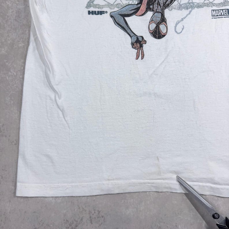 May include: White t-shirt featuring a graphic of Spider-Man in black, red, and gray. The shirt has the "HUF" logo on the left and the "MARVEL" logo on the right. The t-shirt is made of a soft, comfortable material.