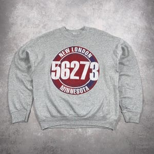 May include: A grey sweatshirt featuring a circular graphic in red, white, and blue. The graphic reads "NEW LONDON 56273 MINNESOTA". The sweatshirt has a crew neck and long sleeves.