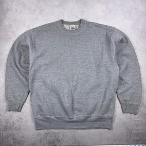 Könnte beinhalten: Ein meliertes graues Sweatshirt mit Rundhalsausschnitt. Das Sweatshirt hat einen gerippten Kragen, Bündchen und Saum. Das Nike-Logo ist auf dem Ärmel sichtbar. Der Stoff scheint ein weiches, bequemes Material zu sein.