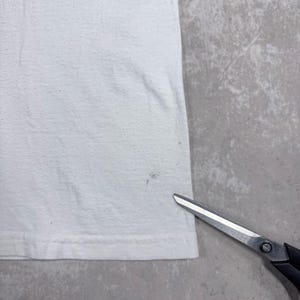 May include: A close-up of a white t-shirt with a small stain near the bottom hem. A pair of scissors with black handles and silver blades is in the lower right corner.