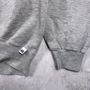 May include: Close-up of a gray sweatshirt sleeve with a ribbed cuff. The Russell Athletic logo is visible on the cuff. The fabric appears to be a soft, medium-weight material. The background is a mottled gray.
