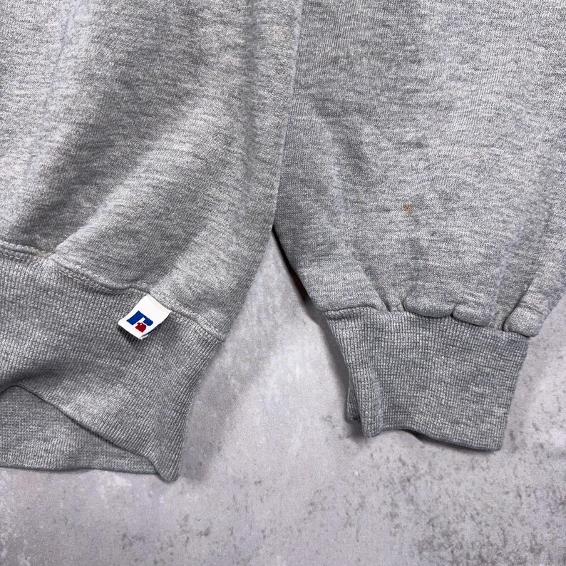 May include: Close-up of a gray sweatshirt sleeve with a ribbed cuff. The Russell Athletic logo is visible on the cuff. The fabric appears to be a soft, medium-weight material. The background is a mottled gray.