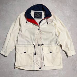 Può includere: Giacca Pacific Trail color crema con cappuccio blu navy e fodera rossa. La giacca ha due tasche frontali, vita con coulisse e chiusura con bottoni. Il logo Pacific Trail è visibile sul cappuccio.